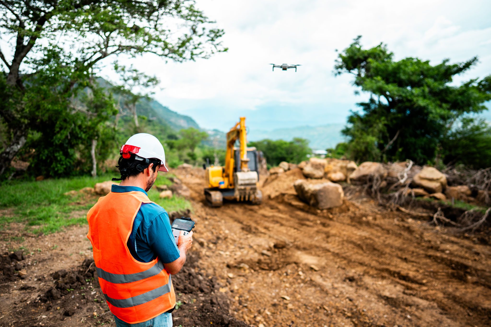 Construction worker monitoring heavy machinery on a dirt road in a mountainous area.